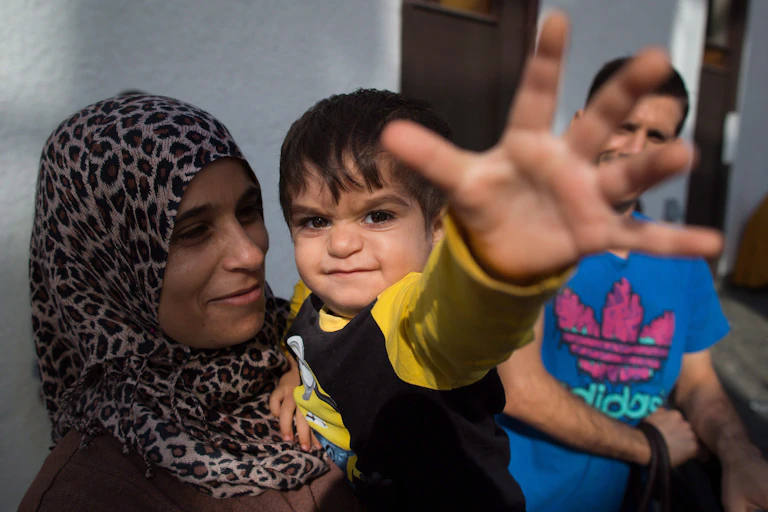 A smiling mother holds her toddler as he reaches for the camera.
