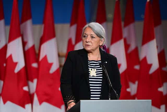 A woman stands in front of a bunch of canada flags behind a podeium