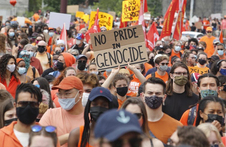 A crowd wearing orange marches down the street, a sign reads 'no pride in genocide'
