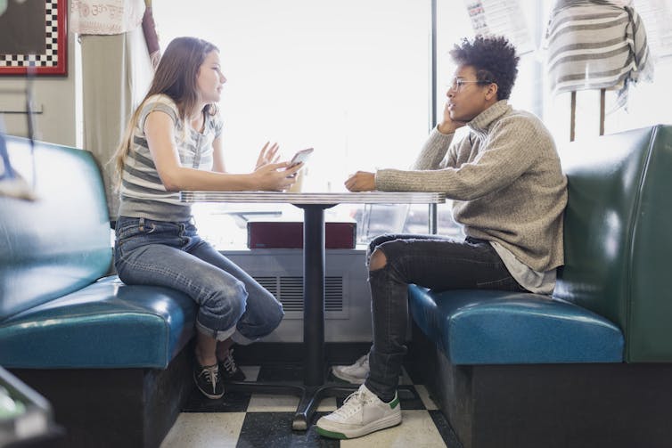 Two teenagers sit across from each other at a table in a cafe. One of them is speaking and the other one is listening.