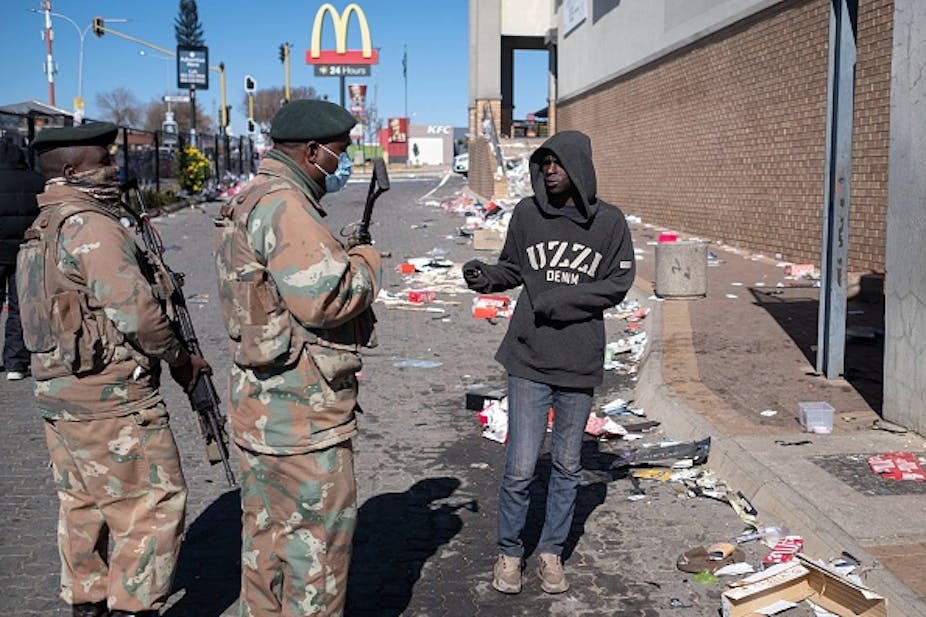 Two armed men in camouflage uniform speak to a civilian outside a large building. Trash scattered on the road.