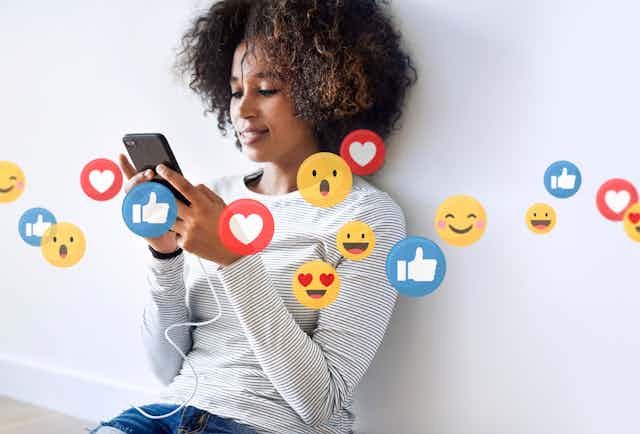 A woman sitting against a wall and smiling at her phone. Images of Like, heart, and smiley emojis float in the air in front of her.