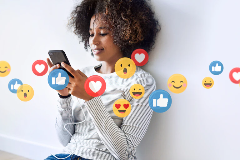 A woman sitting against a wall and smiling at her phone. Images of Like, heart, and smiley emojis float in the air in front of her.