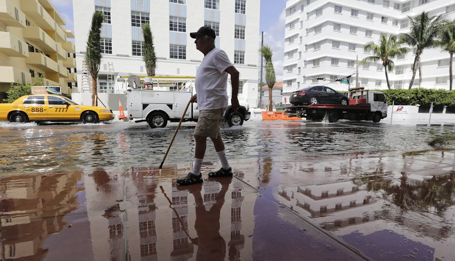 A man with a cane walks on a wet sidewalk next to a flooded street under blue skies