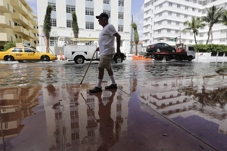 A man with a cane walks on a wet sidewalk next to a flooded street under blue skies