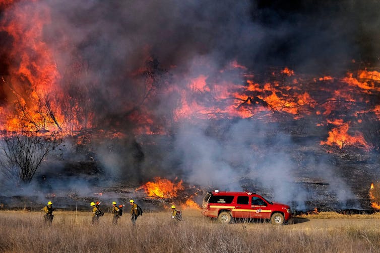 Firefighters and firetruck in foreground, raging fire behind.