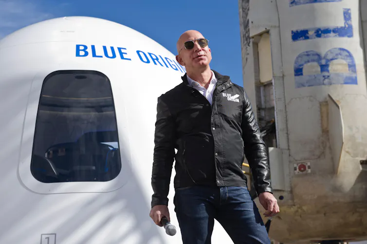 A white man stands in front of a rocket reading 'BLUE ORIGIN'