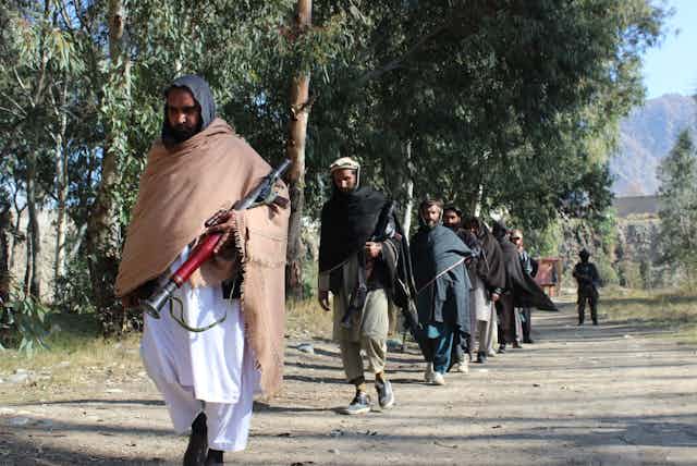 Men in robes with covered heads walk with weapons on a tree-lined dirt road; a soldier can be seen in the background.
