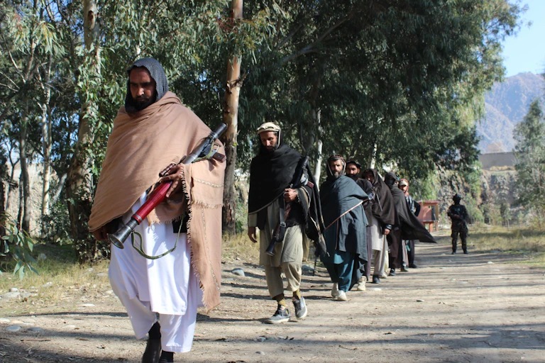 Men in robes with covered heads walk with weapons on a tree-lined dirt road; a soldier can be seen in the background.