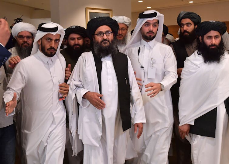 Bearded men in white robes and head coverings walk closely together in a hotel-like setting