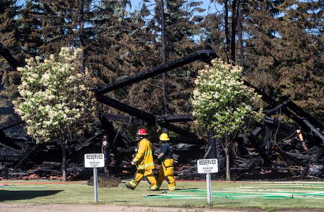 Firefighters walk past the remains of a Catholic church that was on fire, with trees in the foreground and parking signs that read 'reserved for parish staff'