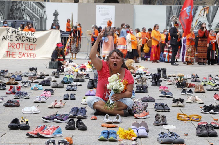 A woman sits with her legs crossed surrounded by hundreds of children's shoes