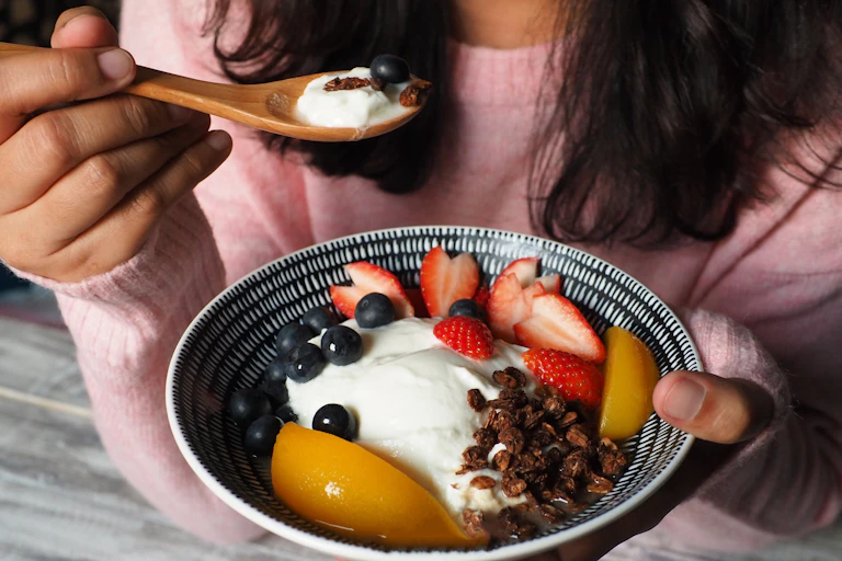 Woman hold bowl of yoghurt and fruit and puts spoon to her mouth.