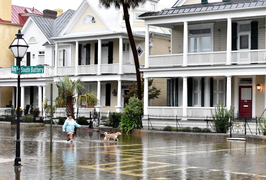 A woman in rain boots walks two dogs through shin-high water in the street past old houses with large porches on South Battery in Charleston