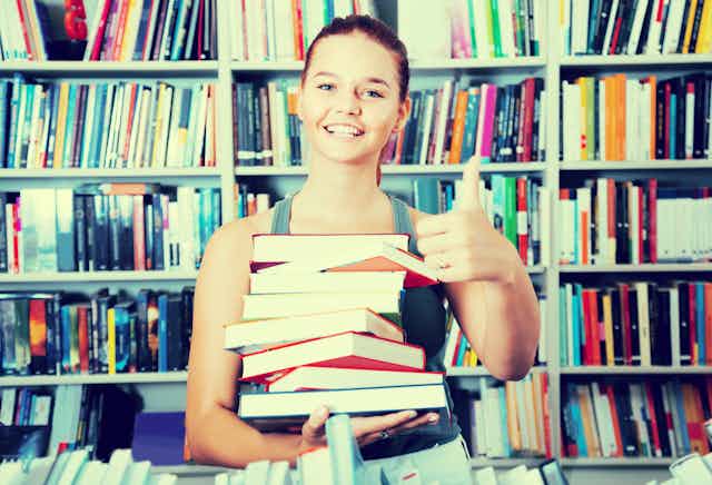 Teen girl holding a stack of books in front of a book shelf.