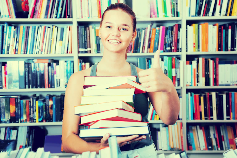 Teen girl holding a stack of books in front of a book shelf.