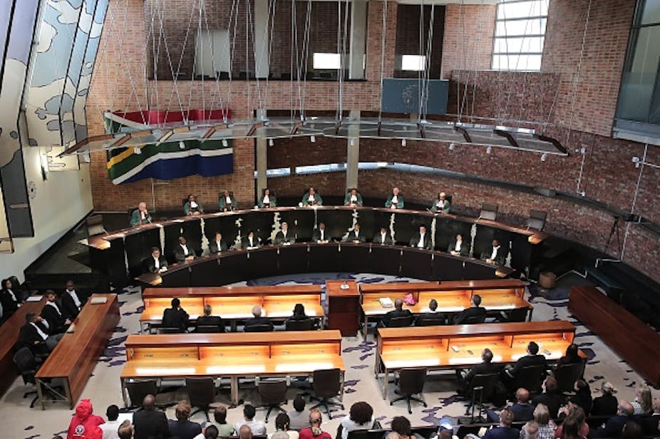 Interior of building, with robed judges sitting at bench facing benches where other people are seated; South African flag on the wall