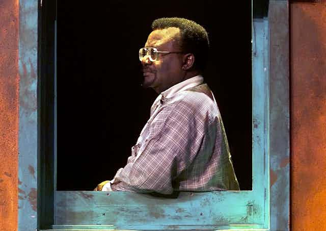 A man in checked shirt stands in profile, leaning his arm on a window frame. He wears glasses and is dramatically lit with what appears to be stage light.