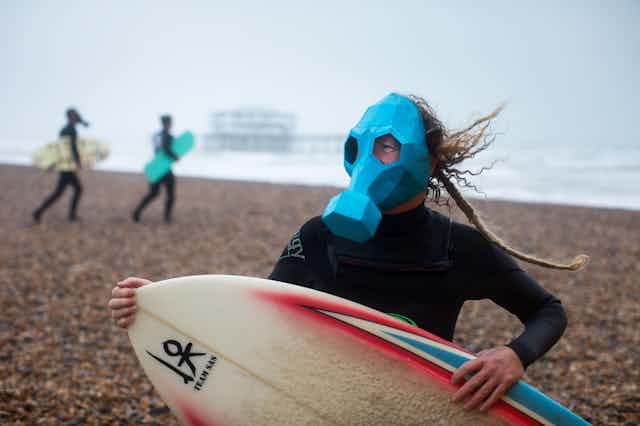 A surfer wearing a gas mask on a shingle beach.