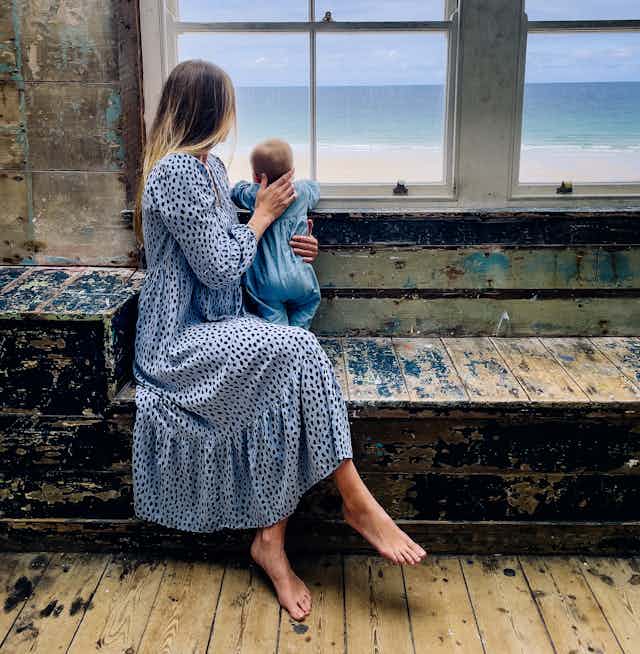 Woman and baby looking out of window towards the sea