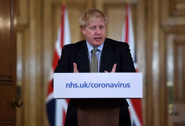 Prime Minister Boris Johnson stands at a podium with a placard reading nhs.uk/coronavirus