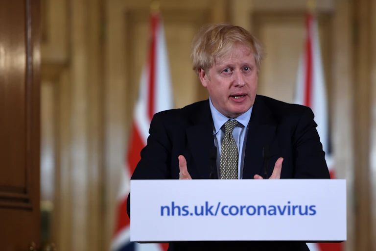 Prime Minister Boris Johnson stands at a podium with a placard reading nhs.uk/coronavirus