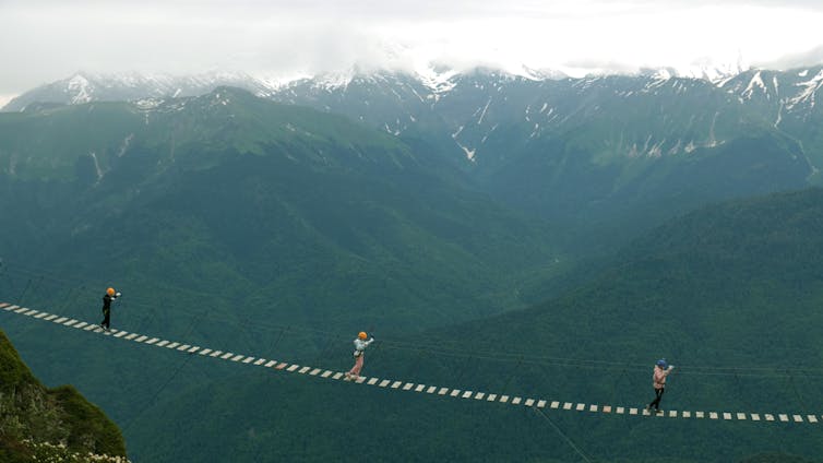 Three people cross a rope bridge against mountain backdrop.
