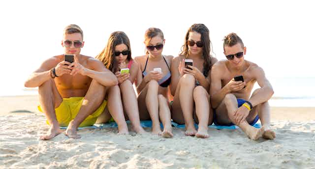 Cinco adolescentes sentados en la playa, en bañador y con gafas de sol, mirando sus teléfonos móviles.