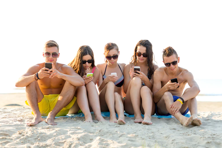 Cinco adolescentes sentados en la playa, en bañador y con gafas de sol, mirando sus teléfonos móviles.