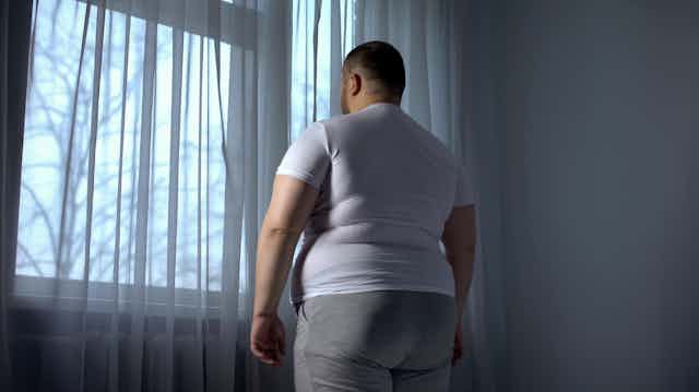 Un hombre con sobrepeso, en una habitación oscura, mira hacia la ventana.
