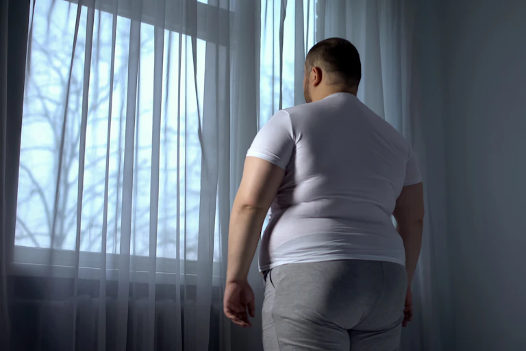 Un hombre con sobrepeso, en una habitación oscura, mira hacia la ventana.