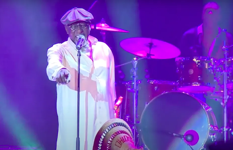 A man on stage in a white shirt and beret, holding a walking stick and pointing to the crowd.