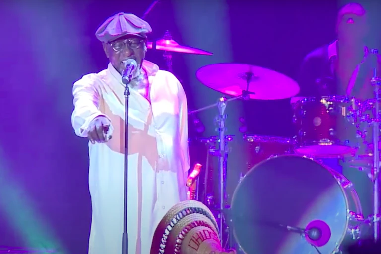 A man on stage in a white shirt and beret, holding a walking stick and pointing to the crowd.