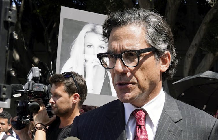 A bespectacled man who is representing Britney Spears as her new lawyer speaks to reporters as a black-and-white photo of Spears hovers in background