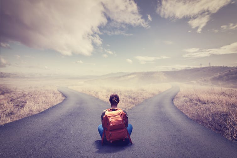 Girl with backpack sitting in front of a road that splits into two.