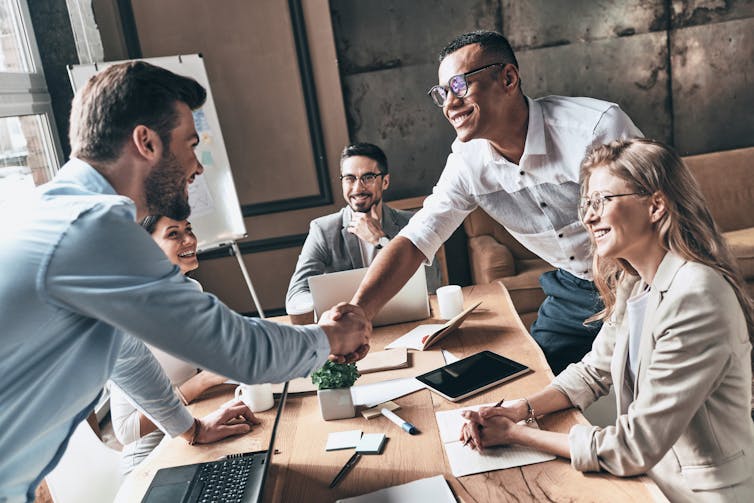 People working together at a desk. New team member reaching over to shake the head of collaborator.