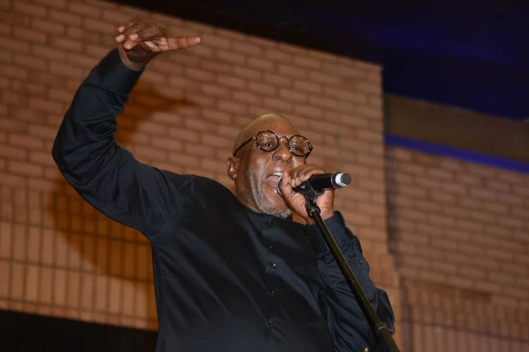 An elderly man on stage singing heartily into a microphone, a hand raised above his head.
