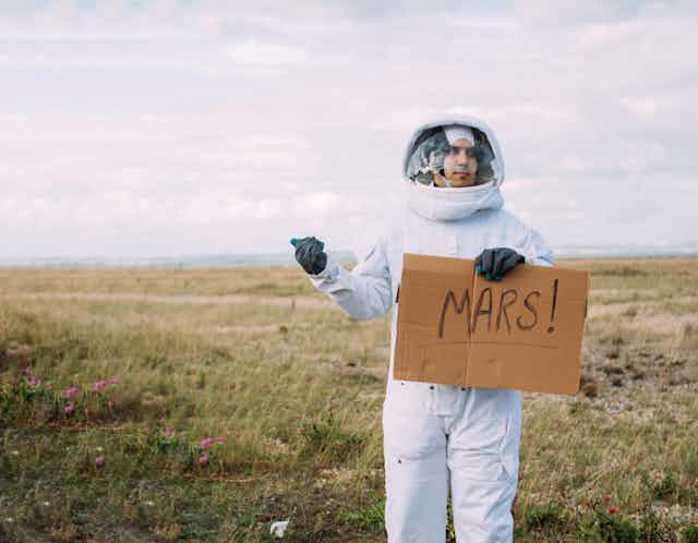 Man in astronaut suit holds up cardboard sign reading 'MARS!' on the side of the road.
