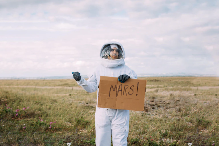 Man in astronaut suit holds up cardboard sign reading 'MARS!' on the side of the road.
