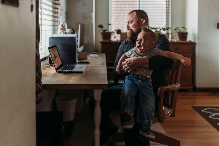 A father holds a crying child on his lap as he works at his computer from home