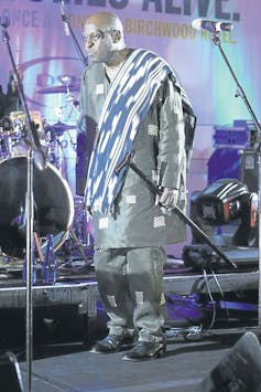 On a live music stage, a balding man in a tunic with cloth over his shoulder holds a walking stick. He is bathed in blue and silver stage light.