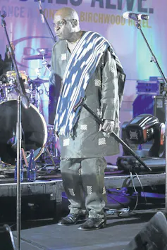 On a live music stage, a balding man in a tunic with cloth over his shoulder holds a walking stick. He is bathed in blue and silver stage light.