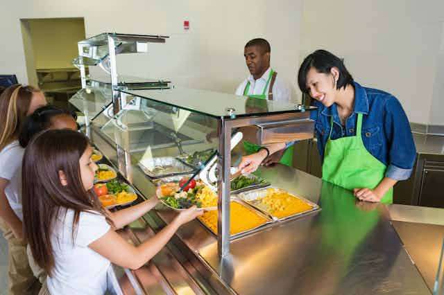 Two cafeteria workers serve food to children during lunch.