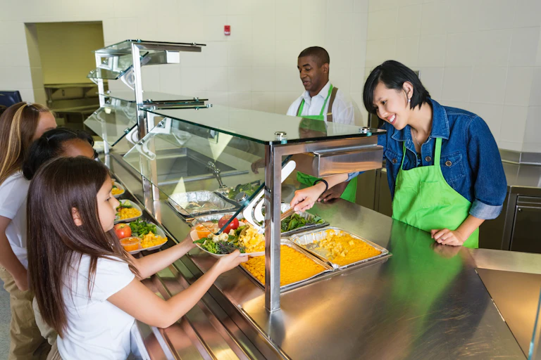 Two cafeteria workers serve food to children during lunch.