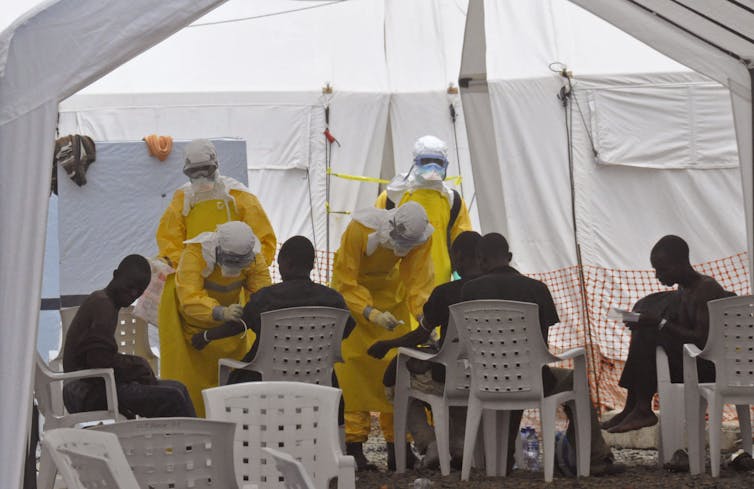 Health care workers in yellow protective equipment treating a group of people in a white tent.