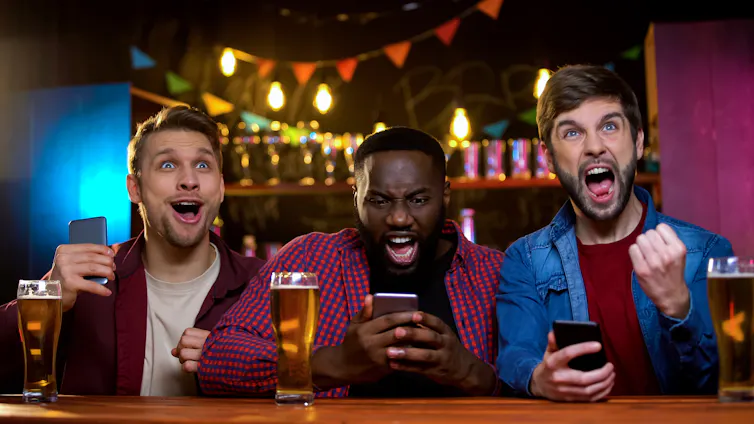 Three sports fans sit at a sports bar with beer, watching the game and looking at their cellphones