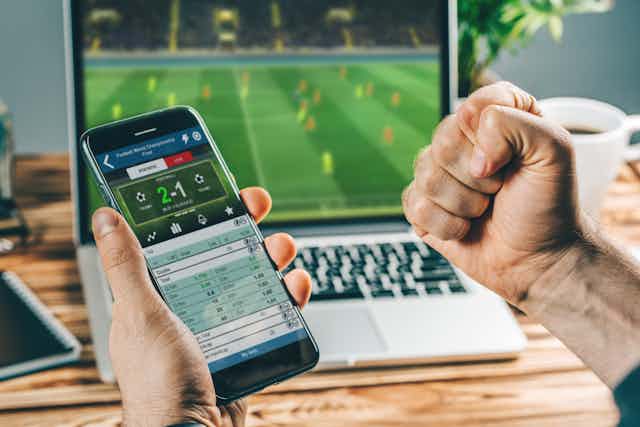 Man sits with his phone in front of a computer, a betting app is open as a soccer match plays in the background