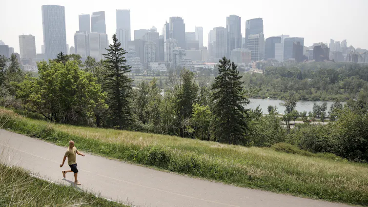 A man walking on a park path with the Calgary skyline in the distance, with a haze of smoke