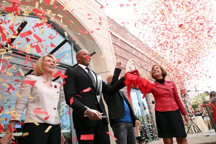 Four people stand before a school building while confetti is blasted around them.