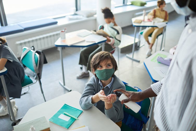 School student wearing mask holds out hands for hand sanitiser offered by masked teacher